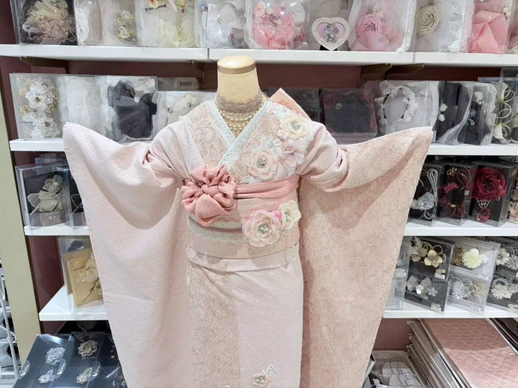 TAKAZENオリジナルのヘップバーンシリーズミニョン。淡いピンク色で、立体のお花が付いているのが特徴的。また、ジャガードとレースの二種類の生地で出来た振袖です。