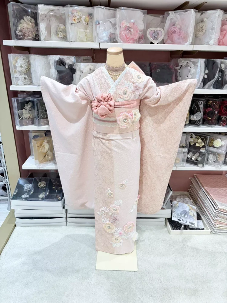 TAKAZENオリジナルのヘップバーンシリーズミニョン。淡いピンク色で、立体のお花が付いているのが特徴的。また、ジャガードとレースの二種類の生地で出来た振袖です。