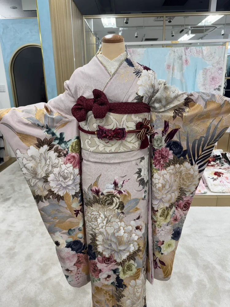 淡いピンクベージュの振袖に白やブルーグレーの牡丹・花柄をあしらった上品な振袖コーデ。金彩が華やかに輝き、ボルドーの帯や帯締め、リボン飾りを合わせた大人可愛い着こなし。