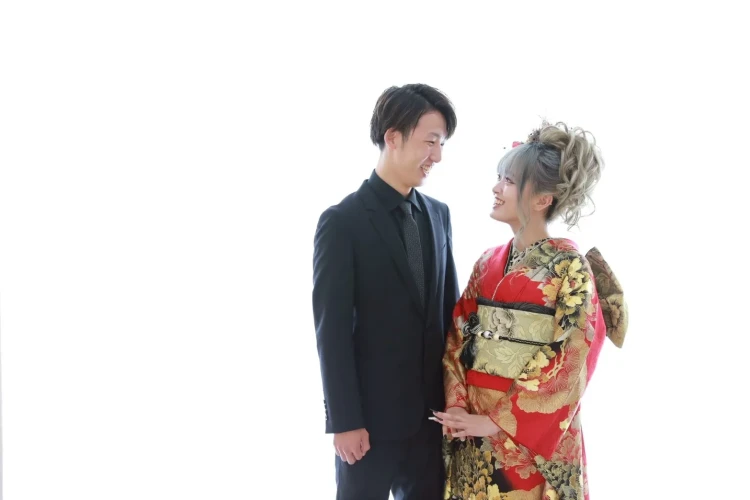 赤い華やかな振袖姿のお嬢様と男性が並んだツーショット。お互いを見つめながら笑顔を見せる、自然で温かみのある記念写真。