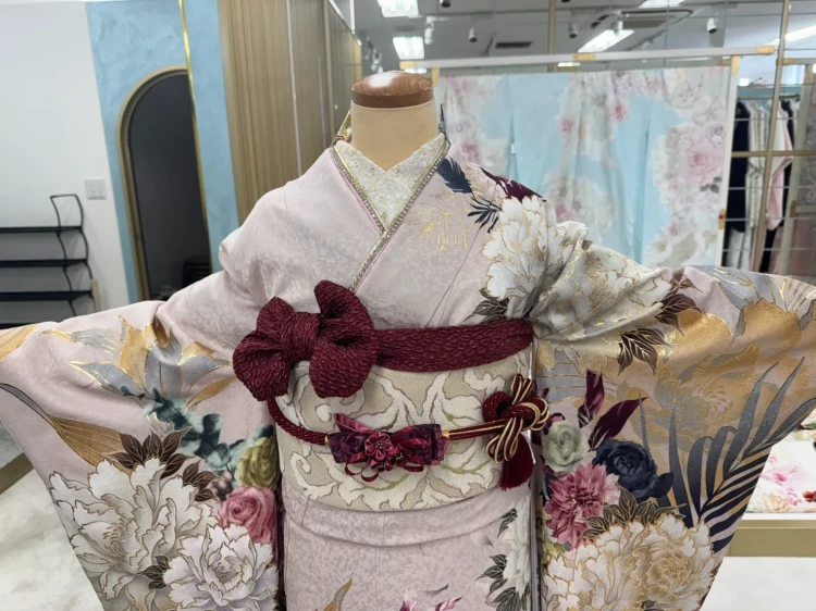 淡いピンクベージュの振袖に白やブルーグレーの牡丹・花柄をあしらった上品な振袖コーデ。金彩が華やかに輝き、ボルドーの帯や帯締め、リボン飾りを合わせた大人可愛い着こなし。