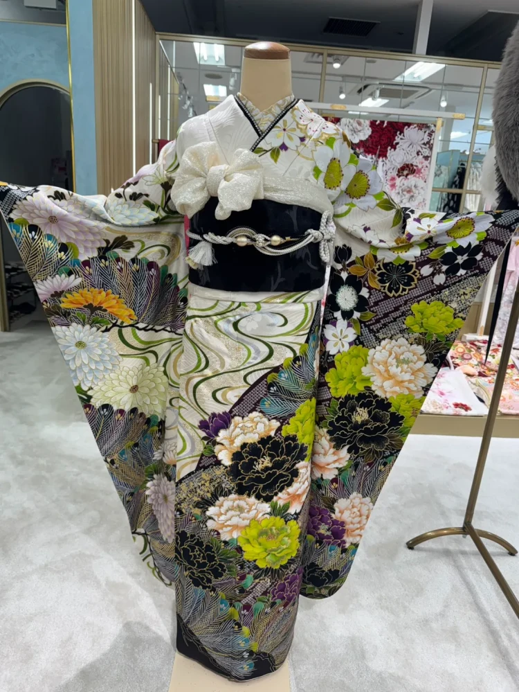 白地に黒・緑・紫の花柄が映える華やかな振袖コーデ。大きな花模様と扇柄が印象的で、黒の帯と白の小物を合わせた大人っぽく個性的なスタイル。